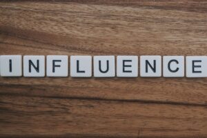 the word "influence" spelled out in scrabble game tiles on a brown wood grain background